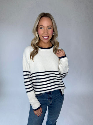 Woman wearing a white sweater with black stripes and blue jeans against a plain background