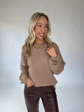 Woman wearing a brown sweater and dark pants against a plain background