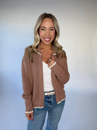 Woman wearing a brown cardigan with white trim, white top, and blue jeans against a plain background