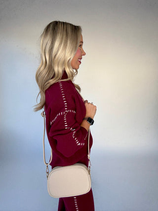 Woman wearing a burgundy sweater with white patterns, holding a beige crossbody bag against a plain background